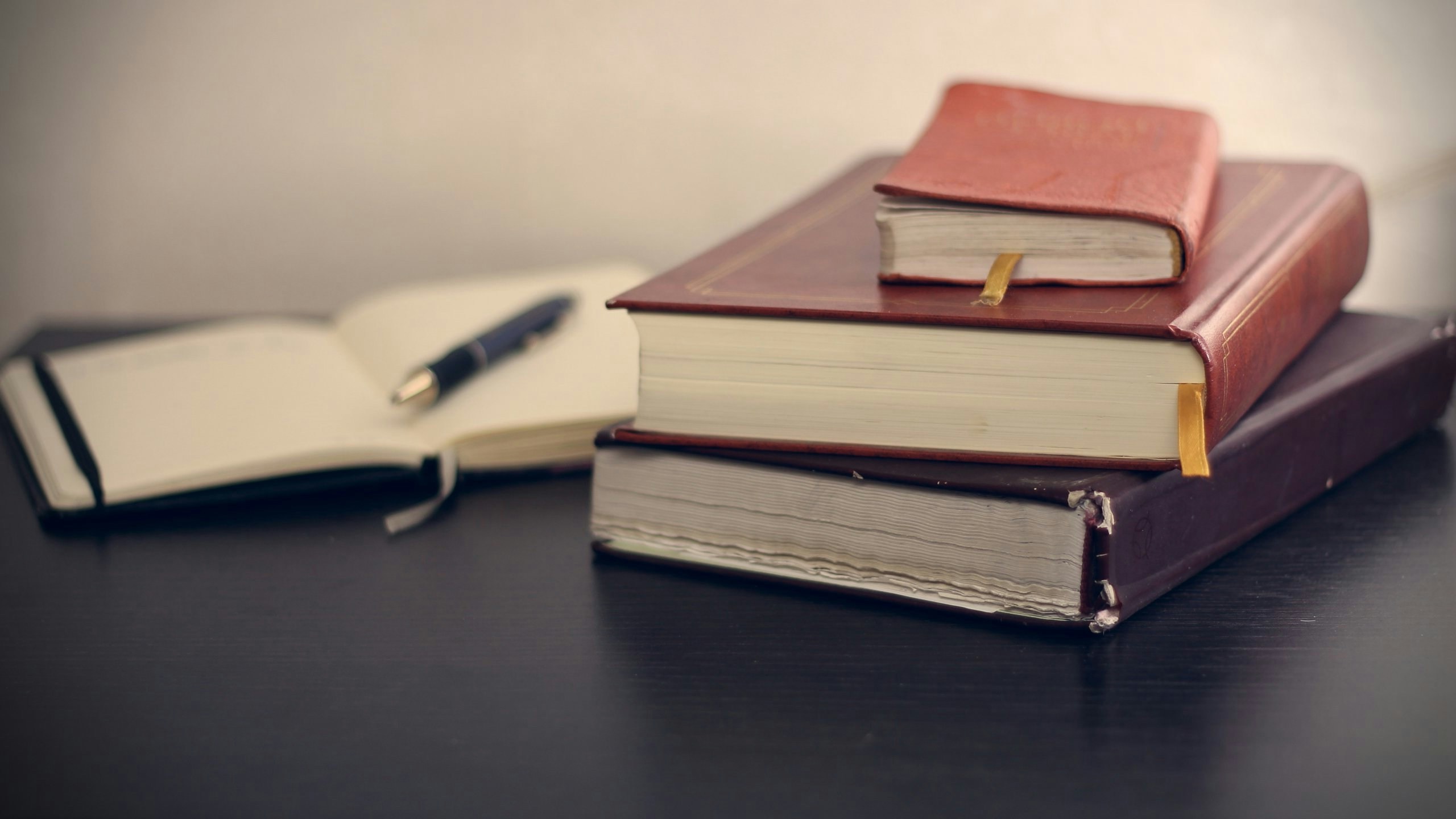 A stack of three leather-bound closed books with a small notebook on top, next to an open notebook and a pen on a dark table.